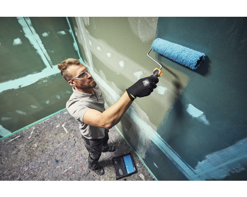 Muž maluje válečkem stěnu v místnosti se sádrokartonovými stěnami a ochrannou rounovou podlahou.