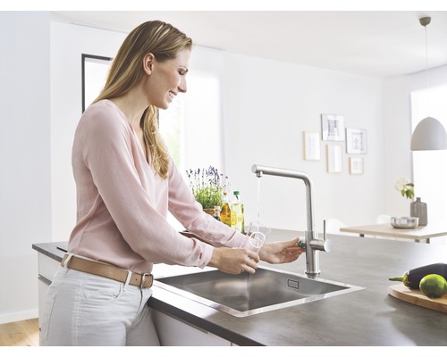 Žena nalévá vodu do sklenice u dřezu s vodovodní baterií ve světlé kuchyni.