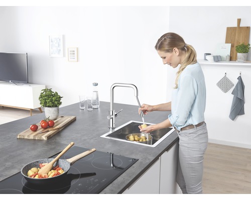 Žena myje brambory v nerezovém dřezu s chromovanou baterií v kuchyni.