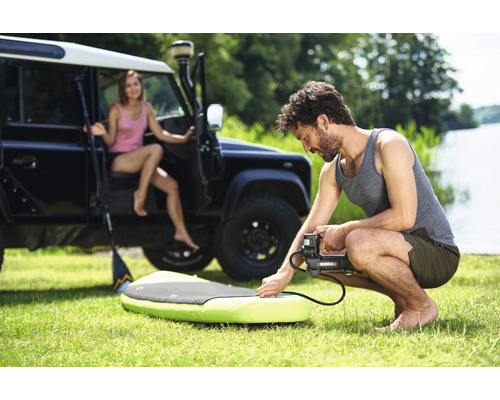 Muž nafukuje paddleboard pomocí elektrické pumpy, v pozadí žena a terénní vozidlo.