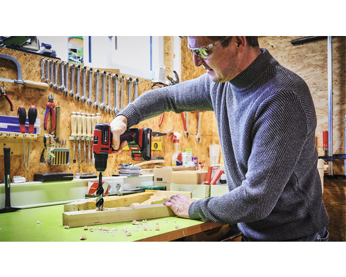 Muž vrtá díru do dřevěné desky akumulátorovým vrtákem v dílně.