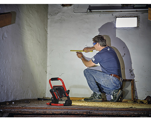 Muž měří stěnu skládacím metrem, prostor osvětluje stavební reflektor
