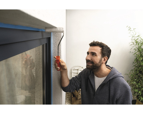 Muž natírá válečkem na barvu rám okna, který je oblepený lepicí páskou.