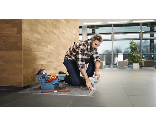 Řemeslník klečí a lepí podlahovou lištu lepicí páskou, vedle je bedna s nářadím a vodováha.