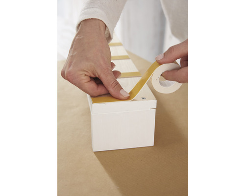 Osoba připevňuje oboustrannou lepicí pásku na bílou dřevěnou krabici.