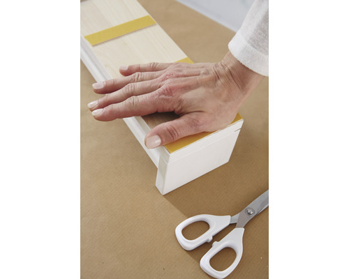 Ruka přitlačuje dřevěnou lištu s lepicí páskou na podklad, vedle leží nůžky.