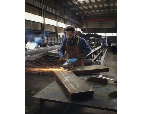 Muž v pracovním oděvu používá v dílně úhlovou brusku na kusu kovu.