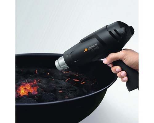 Ruka drží horkovzdušnou pistoli Steinel HL 1821 S nad grilem na dřevěné uhlí.