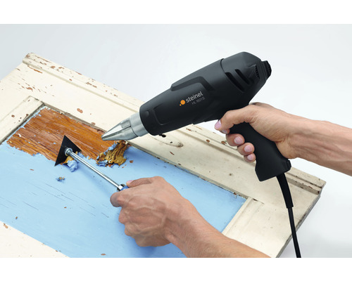 Osoba odstraňuje barvu z okenního rámu pomocí horkovzdušné pistole a škrabky na barvu