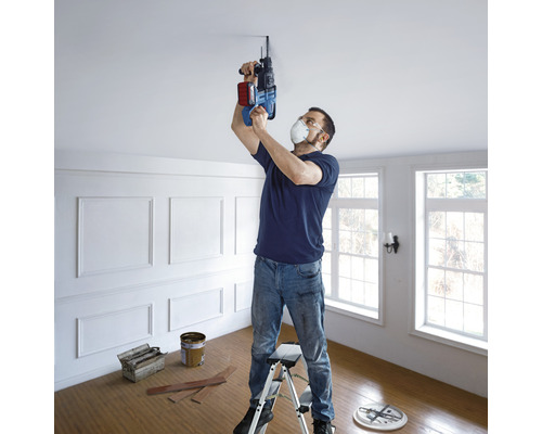 Muž vrtá vrtačkou díru do stropu, stojí na žebříku.