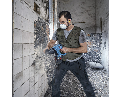 Řemeslník v pracovním oblečení s ochrannými brýlemi a protiprachovou maskou používá na zdi vrtací kladivo.