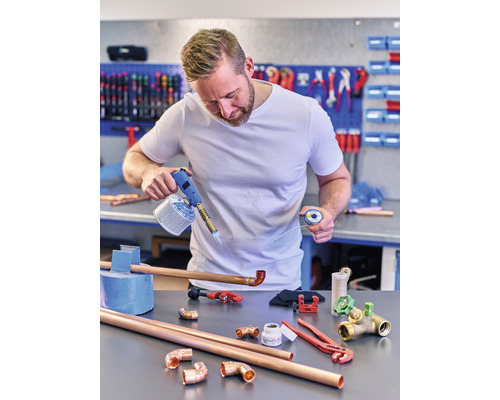 Muž pájí měděné trubky plynovým hořákem a pájkou na pracovním stole s nářadím a tvarovkami.