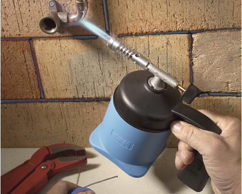 Ruka používá hořák k pájení trubek na cihlové zdi.