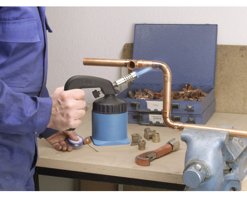 Řemeslník pájí měděné trubky pájecí lampou, kleštěmi na trubky a měděnými tvarovkami na pracovním stole