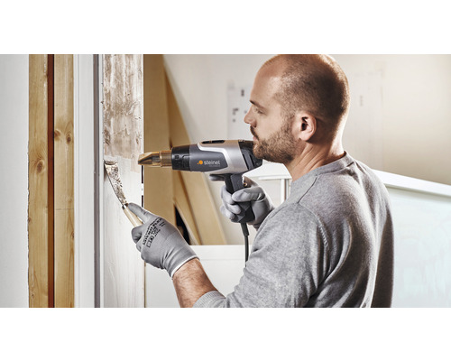 Muž odstraňuje starou barvu z okenního rámu horkovzdušnou pistolí a špachtlí