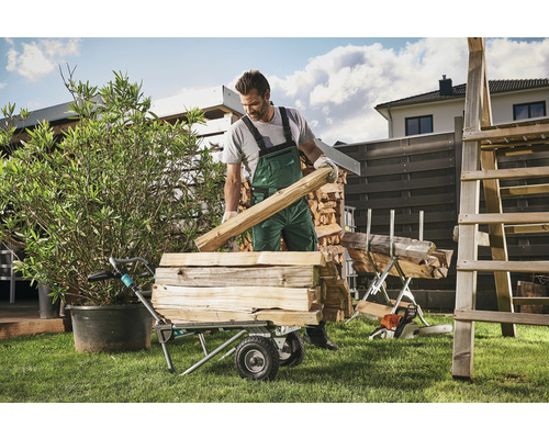 Muž nakládá do transportního vozíku palivové dřevo na zahradě.