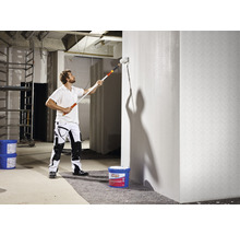 Muž maluje válečkem zeď v hrubé stavbě.