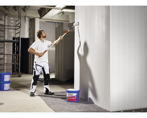 Muž maluje válečkem zeď v hrubé stavbě.