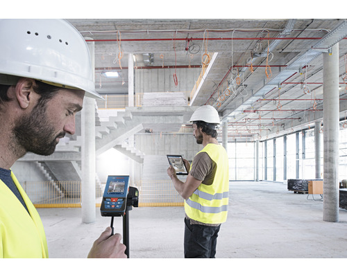 Dva stavební dělníci používají digitální laserový dálkoměr a tablet na staveništi.