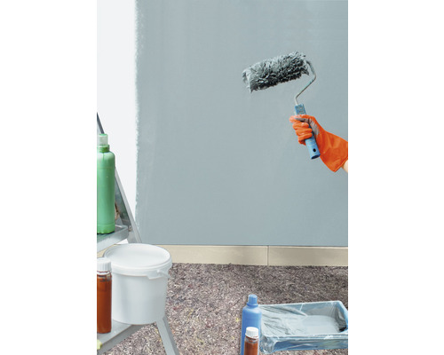 Malování stěny s válečkem, kbelíkem barvy a krycím materiálem na podlaze.