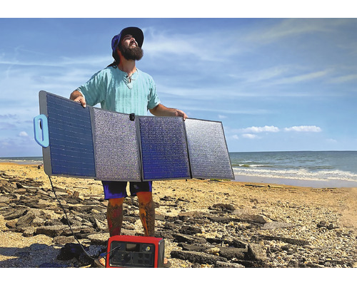 Muž drží skládací solární panelový systém na pláži s napájecí stanicí v popředí.