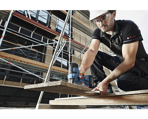 Stavební dělník s ochrannou helmou a brýlemi řeže dřevěné prkno přímočarou pilou na staveništi. V pozadí je lešení.