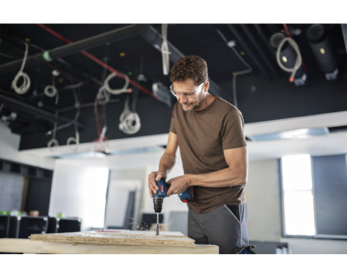 Muž vrtá vrtačkou otvor do dřevěné desky a má na sobě ochranné brýle.