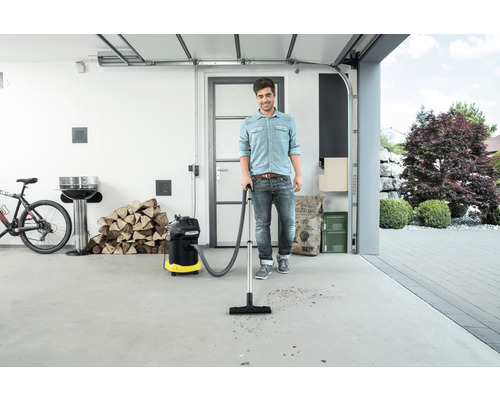 Muž používá víceúčelový vysavač v garáži k odstranění nečistot. V pozadí je vidět gril, dřevo a kolo.