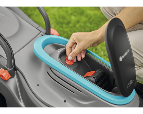 Detailní pohled na sekačku na trávu s otevřenou přihrádkou na baterie a rukou stisknutou na červené tlačítko