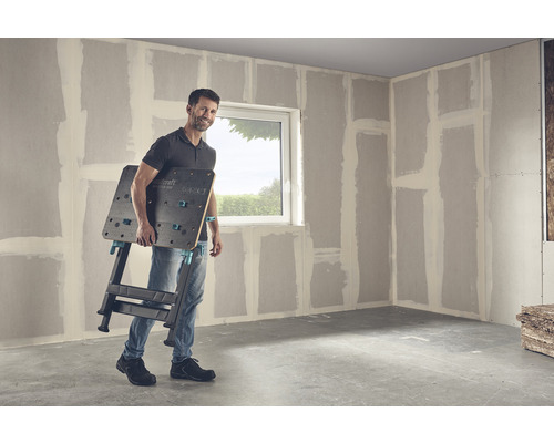 Muž nese pracovní stůl Wolfcraft Master 650 v místnosti se sádrokartonovými stěnami