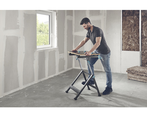 Muž pracuje s pracovní lavicí v místnosti se sádrokartonovými stěnami a oknem.