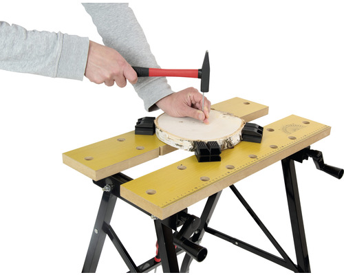 Osoba zatlouká kladivem hřebík do kusu dřeva, který je upevněn pomocí pracovního stolu.