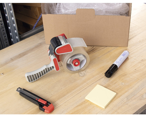 Kartonová krabice, odvíječ lepicí pásky, popisovač, poznámkový blok a řezák na stole.