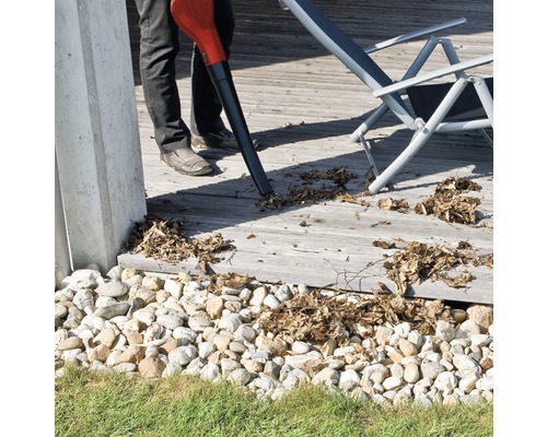 Osoba používá fukar na listí k odstranění listí z dřevěné terasy. V pozadí je vidět lehátko.