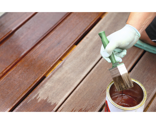 Osoba olejující dřevěnou terasu štětcem a plechovkou oleje.