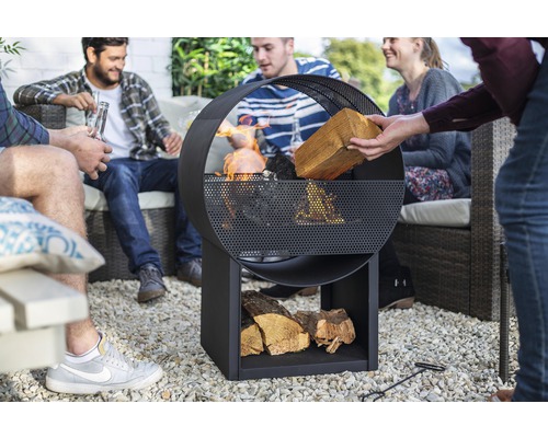 Skupina lidí sedí v zahradě kolem kovového ohniště s úložným prostorem na dřevo.