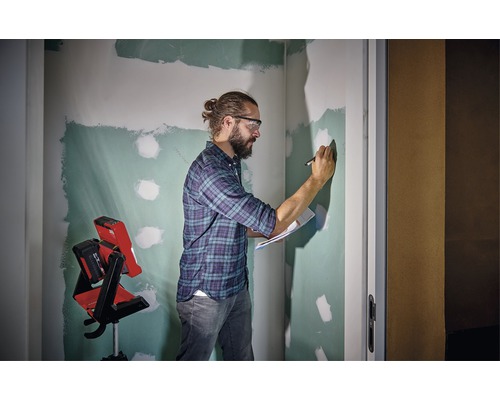 Muž s ochrannými brýlemi, poznámkovým blokem a perem před nedokončenou stěnou a stavebním reflektorem