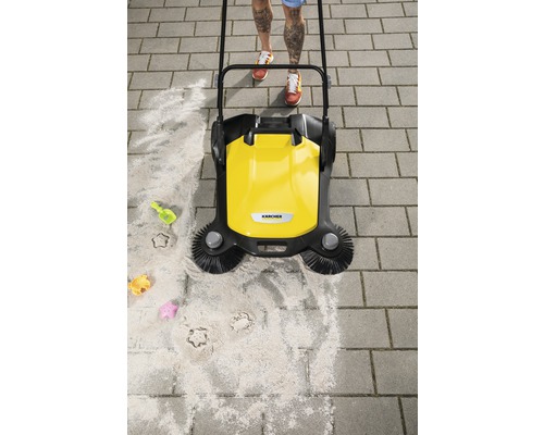 Žlutý zametací stroj v provozu na dlažebních kostkách s pískem a dětskými hračkami.