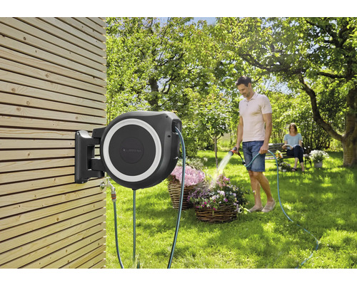 Box na zahradní hadici namontovaná na dřevěné stěně s mužem zalévajícím květiny.