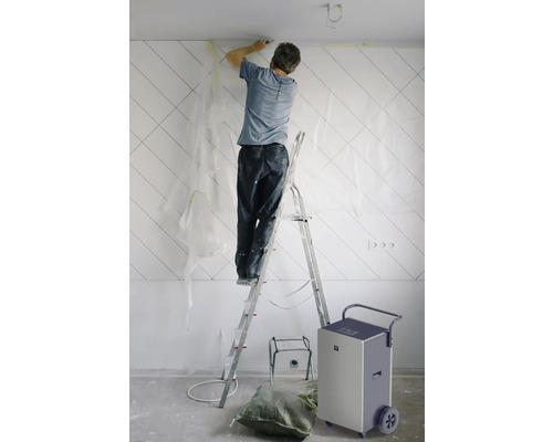 Muž na žebříku renovuje stěnu v místnosti, poblíž je odvlhčovač vzduchu