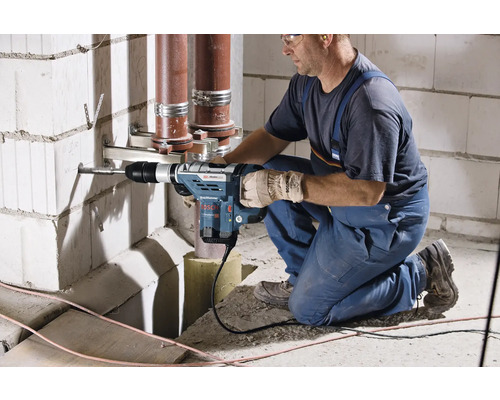 Řemeslník vrtá vrtacím kladivem otvor do zdi, v pozadí vedou trubky.