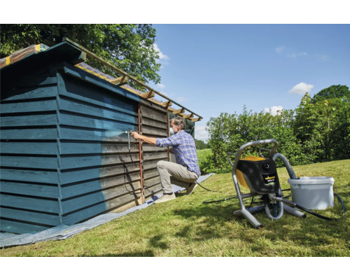 Muž stříká barvou dřevěnou kůlnu pomocí stříkacího systému