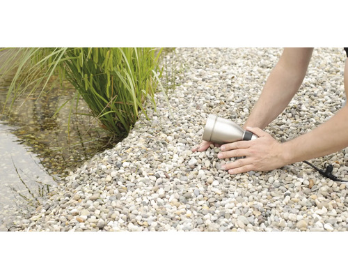 Muž instaluje zahradní reflektor na štěrkové cestě vedle jezírka s rostlinami.