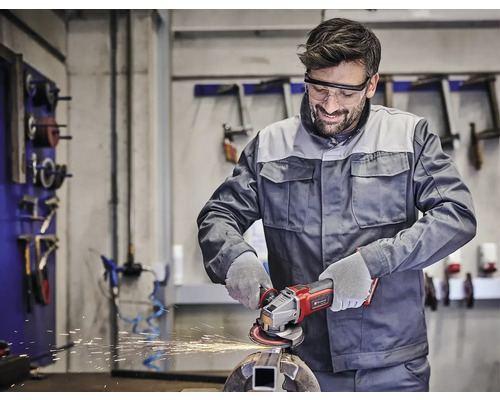 Muž nosí ochranné brýle a pracuje s akumulátorovou úhlovou bruskou na kusu kovu v dílně.