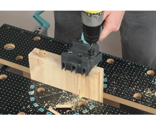 Osoba vrtá vrtačkou díru do kusu dřeva pomocí přípravku pro kolíkování na pracovním stole.