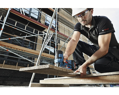 Stavební dělník s helmou a ochrannými brýlemi řeže přímočarou pilou dřevěnou desku na staveništi. V pozadí je vidět lešení. Logo Bosch.