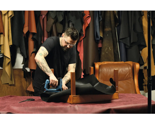 Řemeslník připevňuje elektrickou sponkovačkou černý kus látky k dřevěnému rámu v dílně.