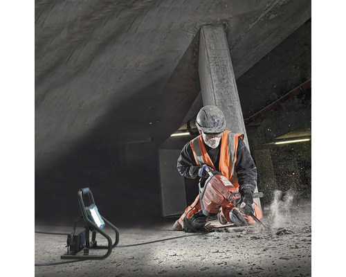 Stavební dělník používá bourací kladivo pod betonovou konstrukcí, osvětlené pracovním světlem.