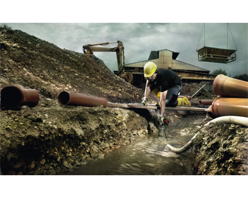 Stavební dělník pracuje v příkopu s trubkami, bagrem a jeřábem v pozadí.