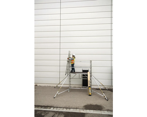 Osoba s ochrannou přilbou a bezpečnostní vestou stojí na pojízdném lešení a drží žebřík u zdi.